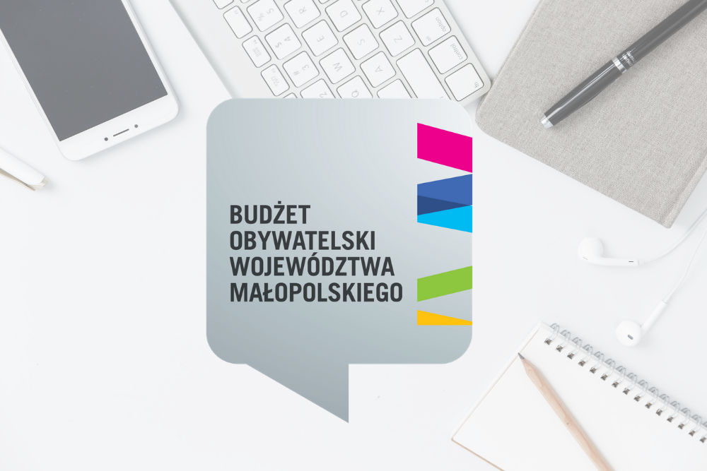 Po środku obrazka znajduje się chmurka, w której widnieje napis Budżet Obywatelski Województwa Małopolskiego. W chmurce z prawej strony znjaduje się patren z loga Małopolski. Do okoła obrazka są także: biały telefon, biała klawiatura,szary notes z czarnym długopisem, białe słuchawki oraz notes z ołówkiem.